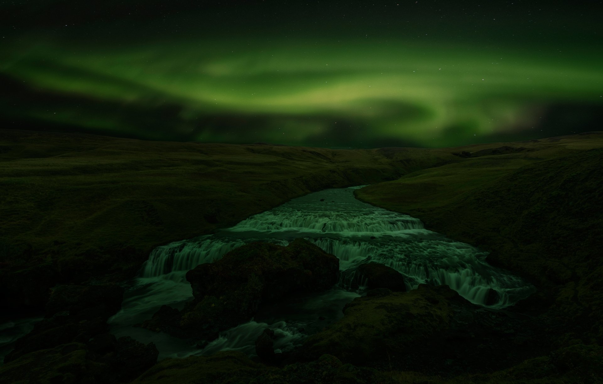 8K Ultra HD PC desktop wallpaper/background — night nature scene: vivid green aurora borealis swirling above a dark river and rugged terrain.
