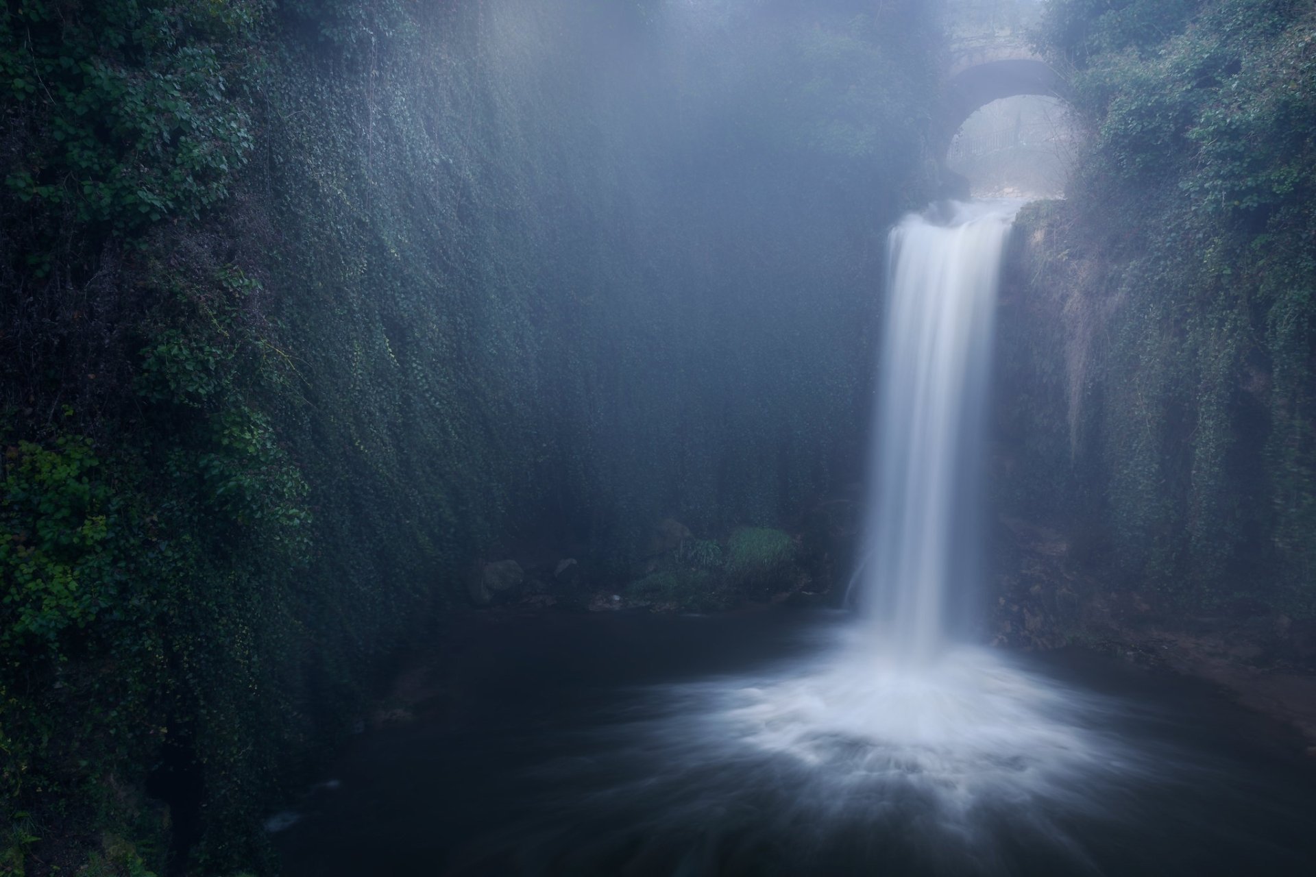 HD desktop wallpaper featuring a serene waterfall cascading into a misty pool, surrounded by lush greenery and rocky cliffs in a tranquil natural setting.