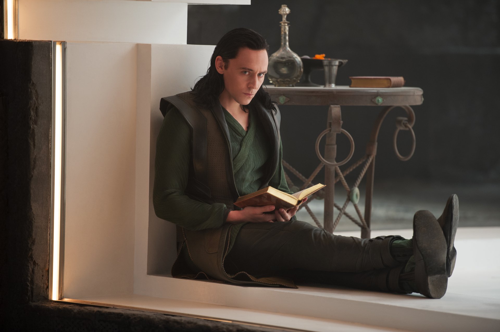Loki from Thor: The Dark World sits against a ledge reading a book in a moody movie still, presented as a 5K Ultra HD PC desktop wallpaper and background.