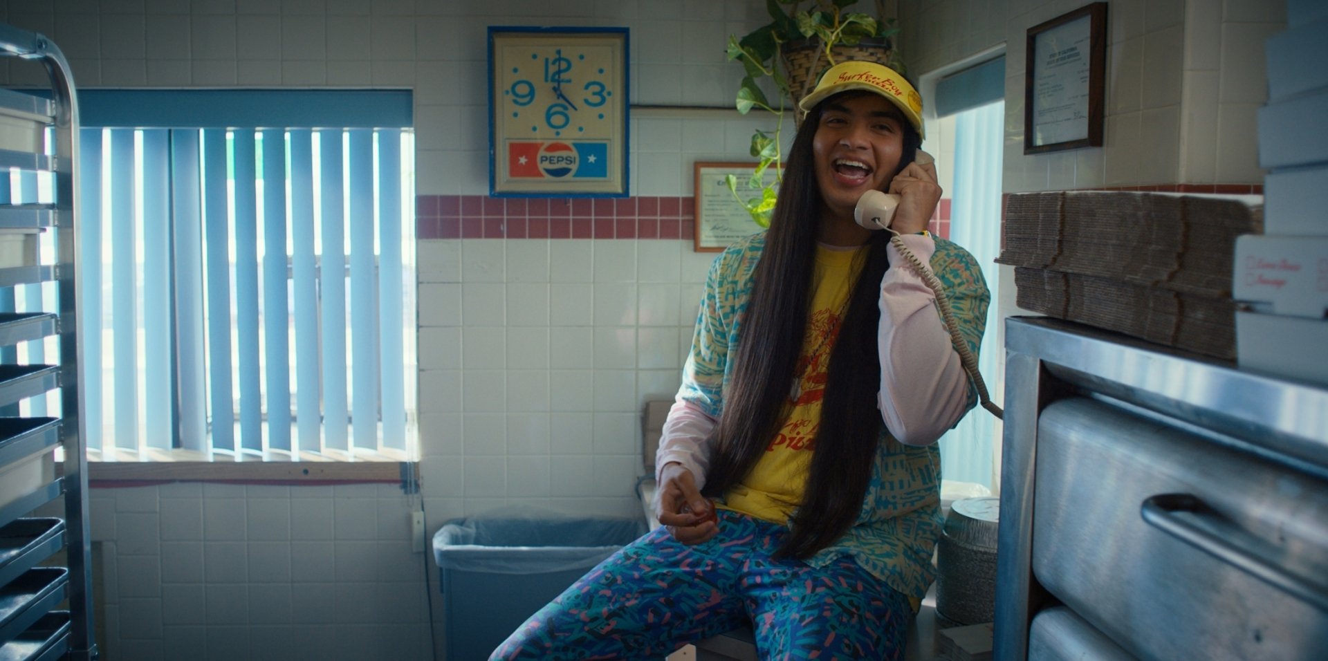 HD PC desktop wallpaper of a cheerful character from the TV show Stranger Things, sitting in a diner phone booth wearing bright 80s clothes and a yellow cap, laughing on the phone.