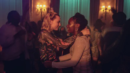 Two women embrace and smile on a warmly lit dance floor — Heartstopper TV show, 4K Ultra HD PC desktop wallpaper.