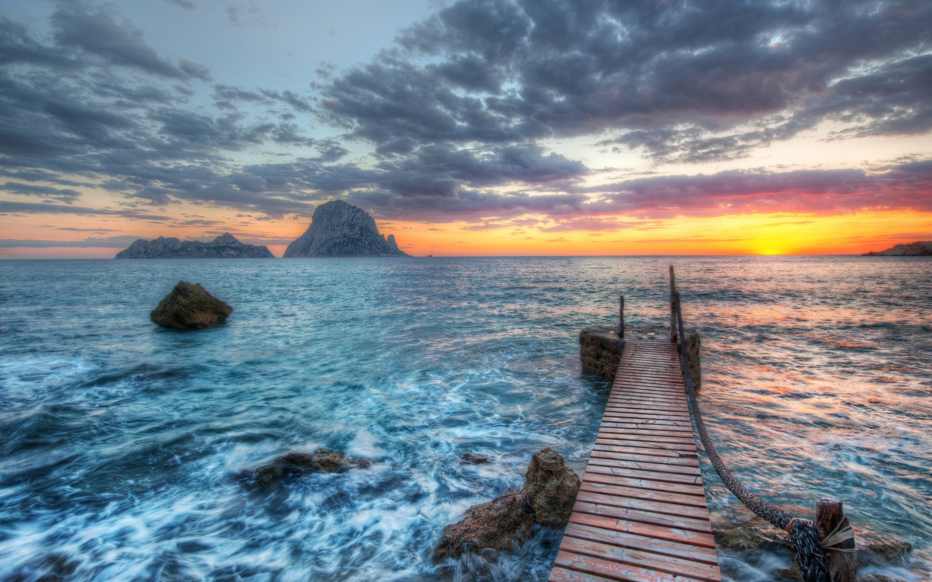 HD PC desktop wallpaper showing a wooden pier extending into a vibrant ocean scene with rocky formations under a dramatic cloudy sunset sky.