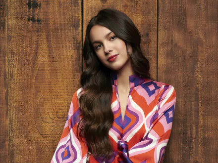 Olivia Rodrigo poses against a wooden backdrop in a colorful patterned outfit, featured as an HD desktop wallpaper from High School Musical: The Musical: The Series.