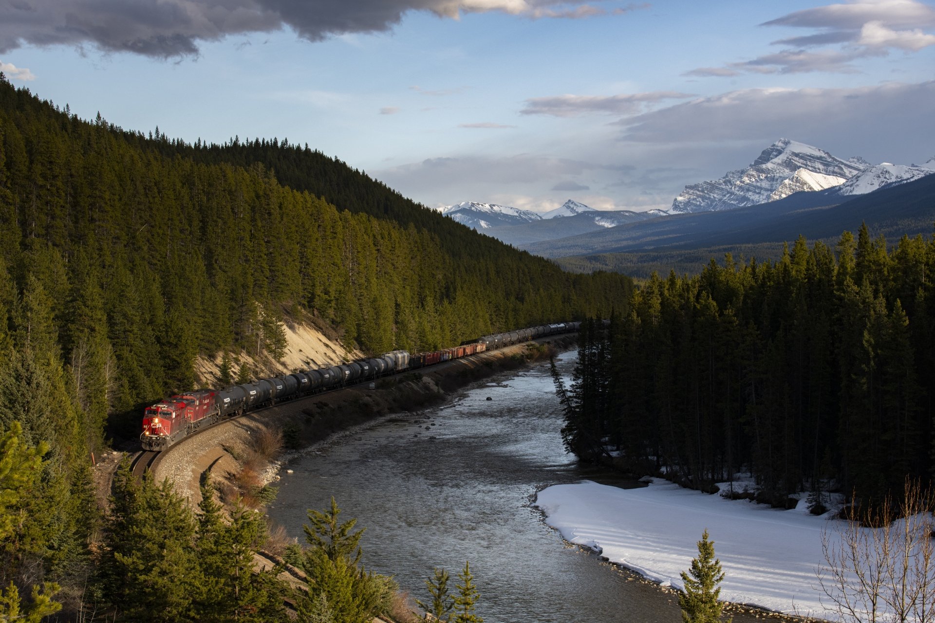 8K Ultra HD PC desktop wallpaper: a red freight train (vehicle) curving along a river through snow-dusted mountains and dense pine forest.
