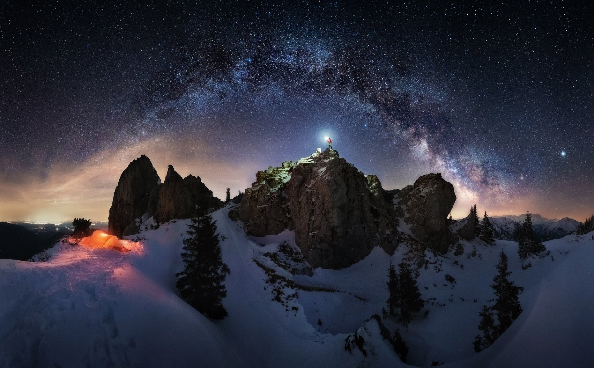 HD desktop wallpaper capturing a starry night sky over snowy mountains with a camper's light glowing beneath the Milky Way, blending nature and night photography.