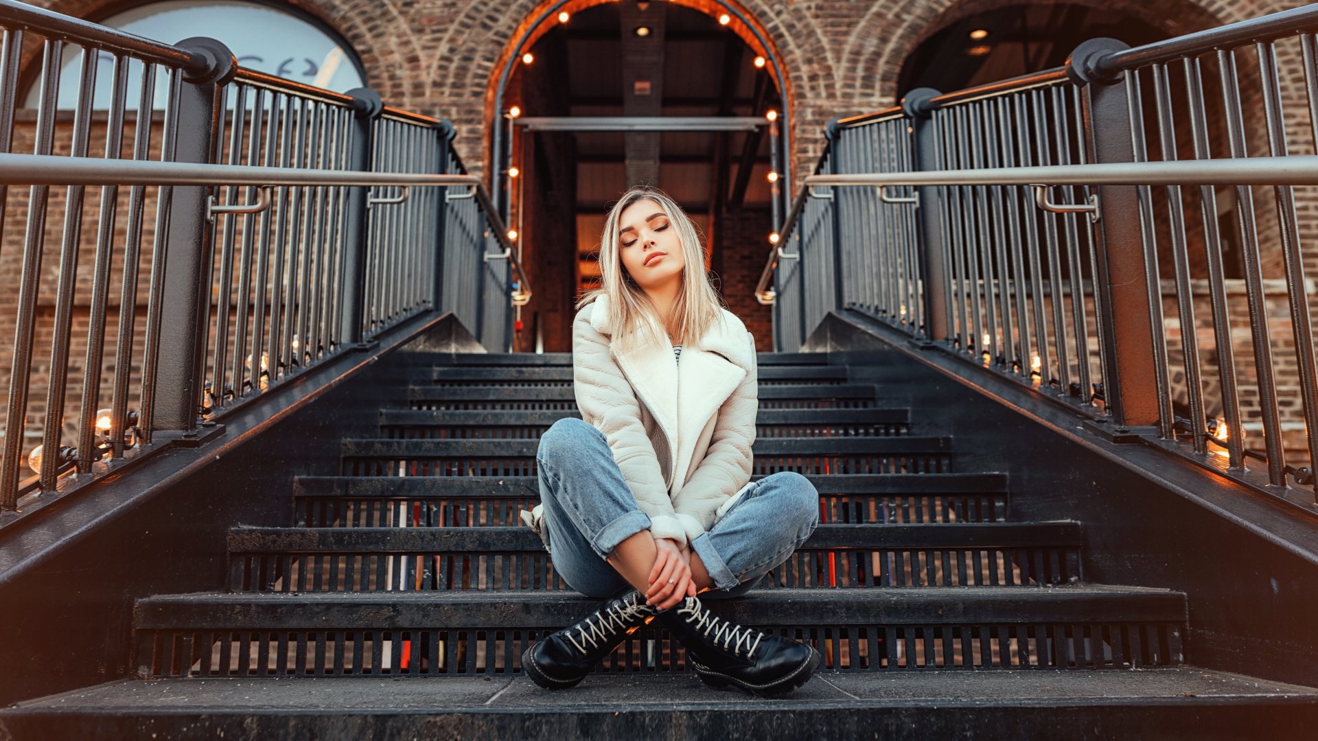 8K Ultra HD PC desktop wallpaper and background of a woman model seated on urban metal stairs under warm string lights, wearing a white jacket and jeans, looking at the camera.