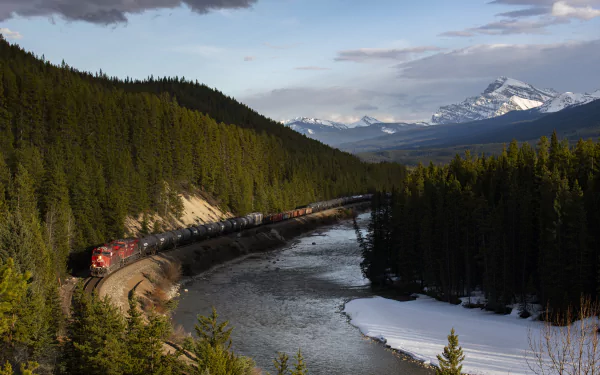 8K Ultra HD PC desktop wallpaper: a red freight train (vehicle) curving along a river through snow-dusted mountains and dense pine forest.