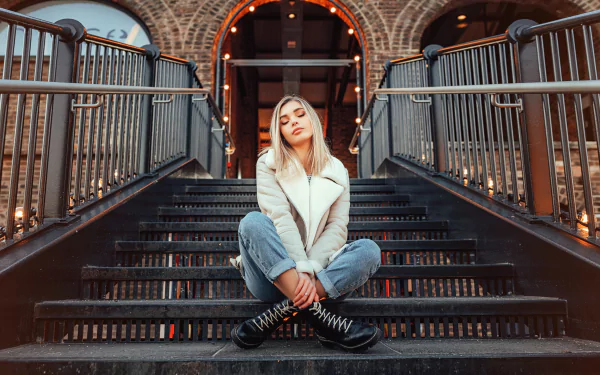8K Ultra HD PC desktop wallpaper and background of a woman model seated on urban metal stairs under warm string lights, wearing a white jacket and jeans, looking at the camera.