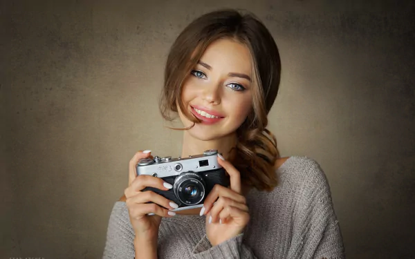 HD PC desktop wallpaper: smiling woman model in a cozy sweater holding a vintage camera against a warm, textured backdrop.
