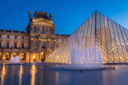 Evening view of the Louvre Museum and glass pyramid in Paris, France, with illuminated fountains reflecting on the water.