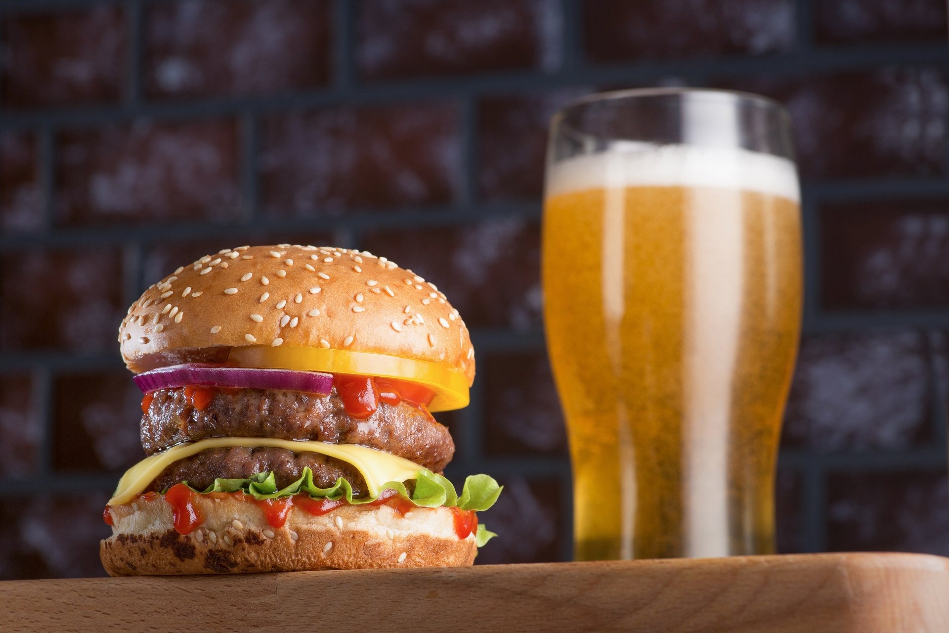 Food, burger: juicy cheeseburger with lettuce, tomato and sauce next to a frosty glass of beer on a wooden surface; 2K Quad HD PC desktop wallpaper and background with brick wall backdrop.