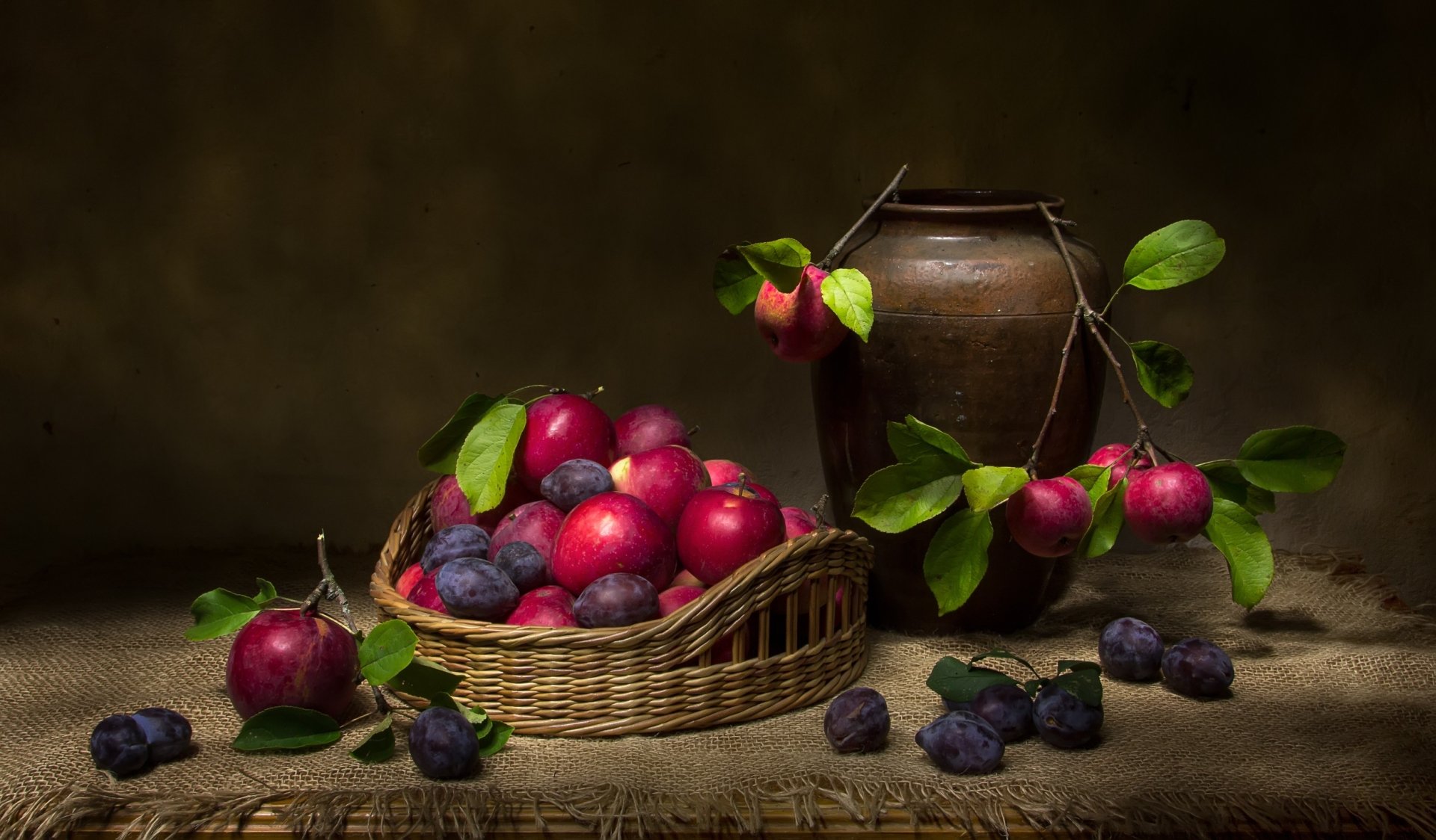 HD PC desktop wallpaper: still life of plums — ripe fruit in a wicker basket and scattered on a rustic table beside an earthenware jug, accented by green leaves and soft lighting.