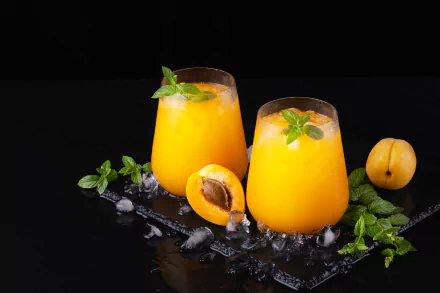 HD PC desktop wallpaper featuring two glasses of vibrant orange juice garnished with mint, surrounded by ice cubes and fresh apricots on a black background.