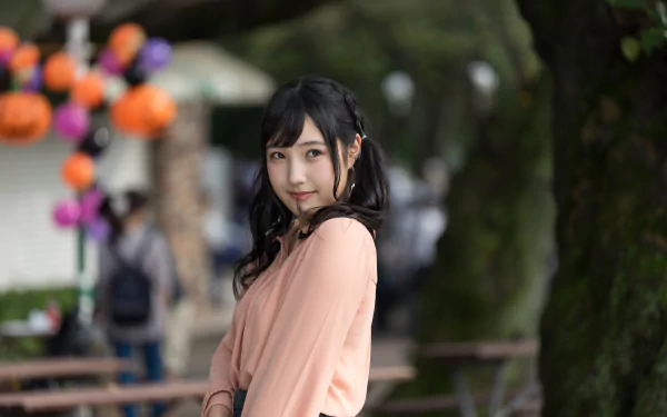 Asian woman with dark hair in a soft pink top, standing outdoors with blurred trees and festive decorations in the background, captured in 4K Ultra HD quality.