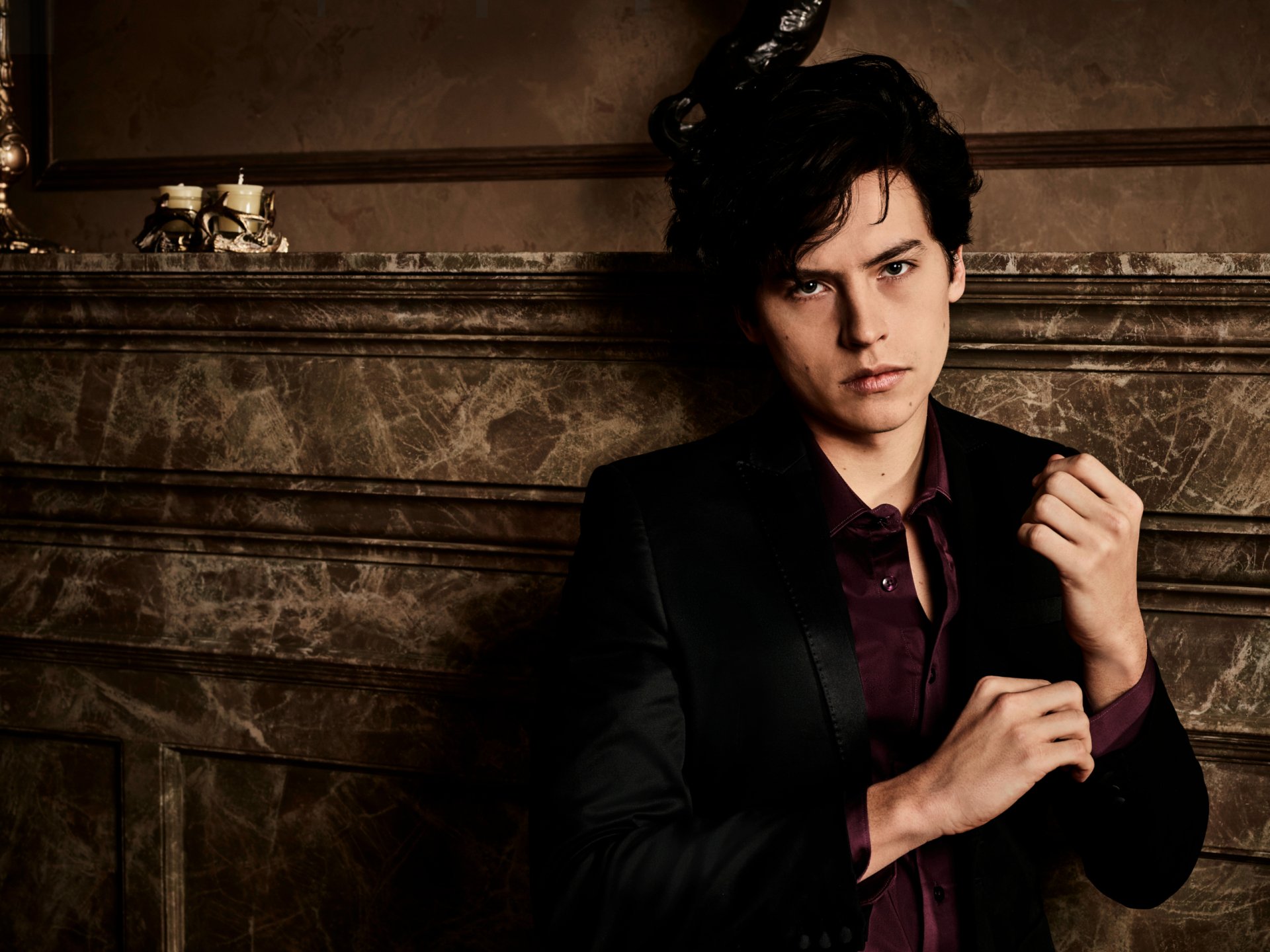 Brooding young man in a dark suit before a marble fireplace, adjusting his cuff — TV show Riverdale, 2K Quad HD PC desktop wallpaper/background.