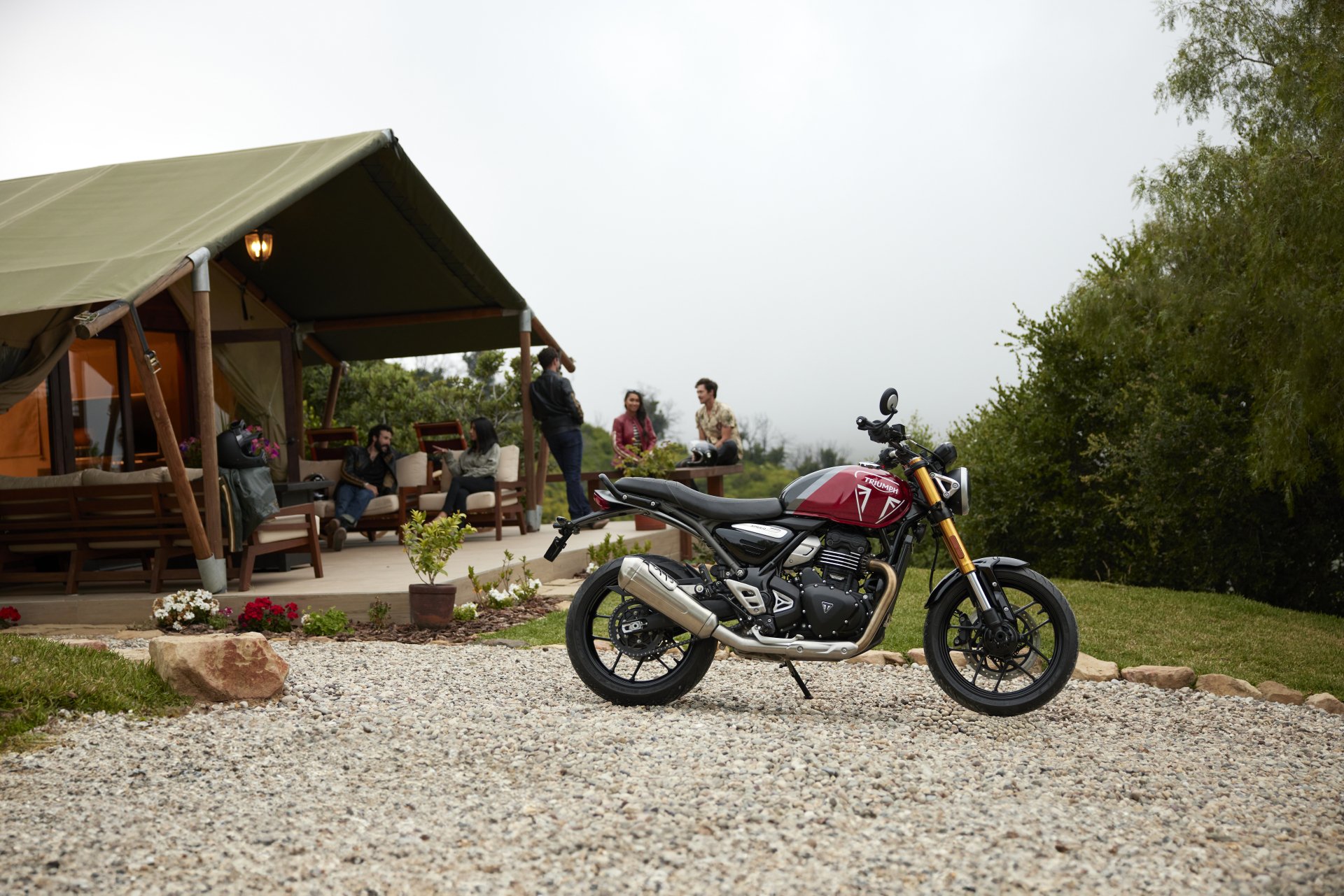 HD wallpaper of a Triumph Speed Twin 400 motorcycle parked in front of a cozy tent with people in the background.