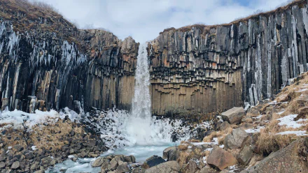 4K Ultra HD wallpaper of Svartifoss waterfall cascading amid distinctive basalt columns and rocky terrain under a cloudy sky in a natural Icelandic landscape.
