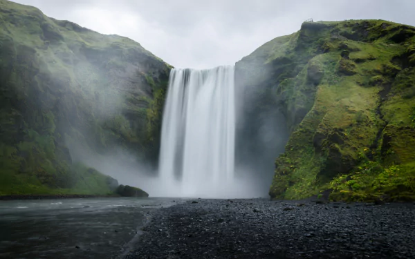 4K Ultra HD image of the majestic Skógafoss waterfall cascading between lush green cliffs under a cloudy sky, capturing the raw beauty of nature.