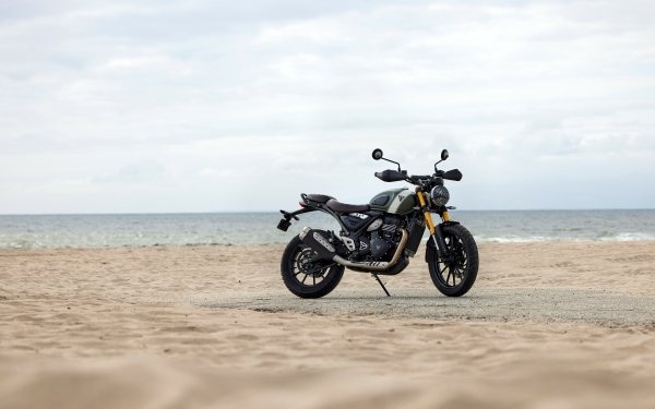 HD wallpaper of a Triumph Scrambler 400 X motorcycle parked on a sandy beach with overcast skies in the background.
