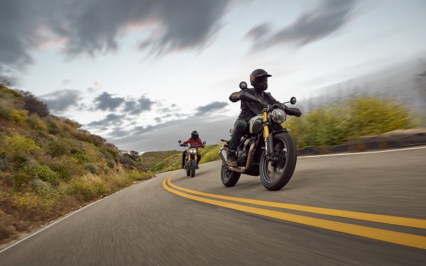 Two riders on Triumph Scrambler 400 X motorcycles speeding along a winding road in a dynamic HD desktop wallpaper background.