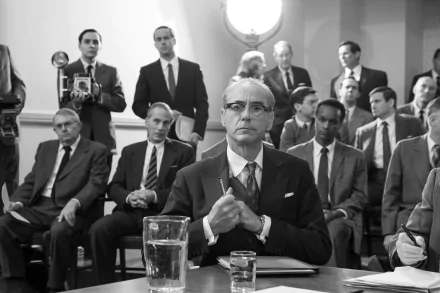A striking black-and-white scene from the movie Oppenheimer, featuring Robert Downey Jr. seated at a table, surrounded by a crowd of onlookers, capturing a tense moment.