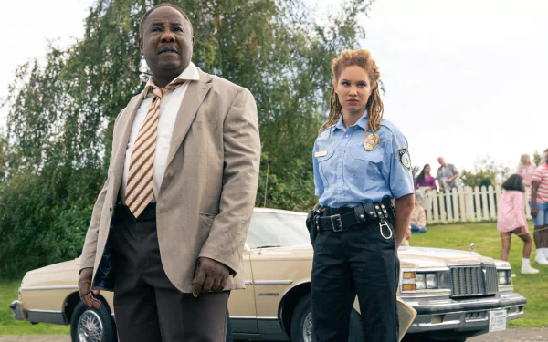 HD wallpaper featuring two actors portraying police officers standing confidently beside a vintage car, set against a grassy outdoor backdrop, associated with the movie 'Cocaine Bear'.