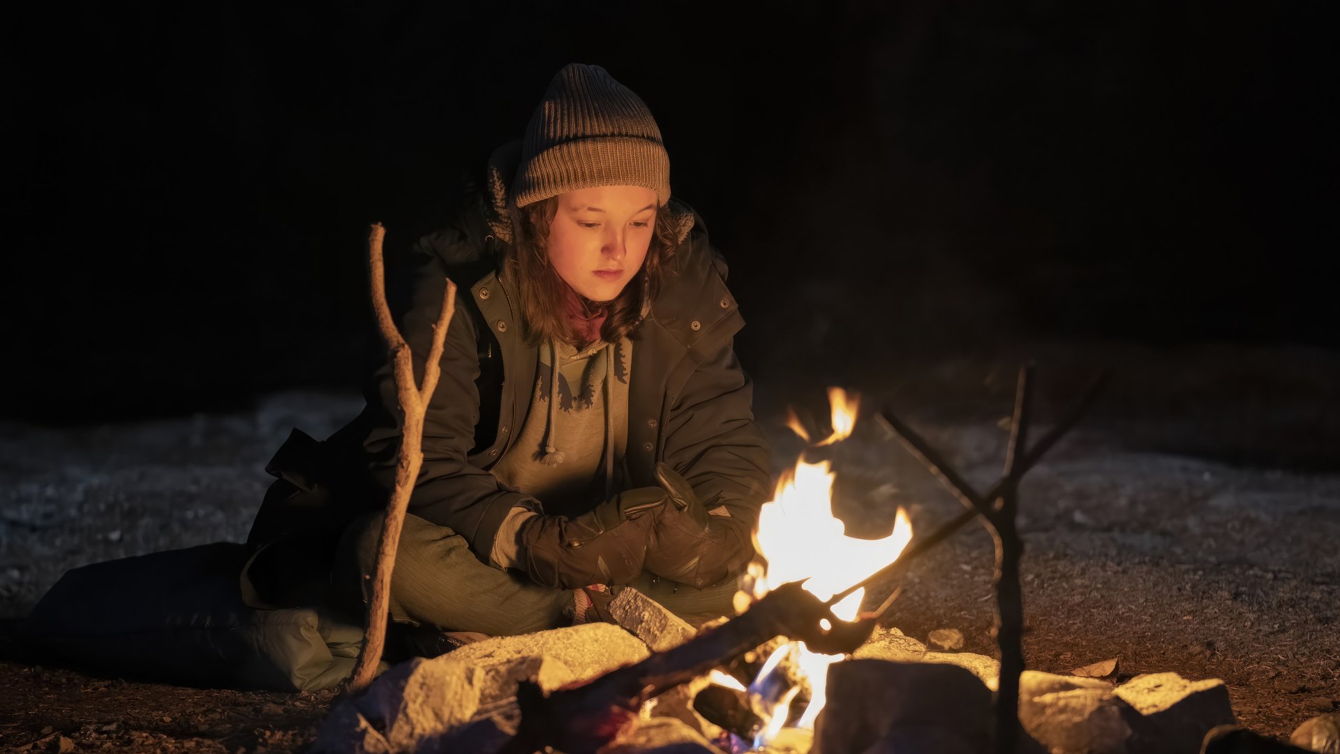 Bella Ramsey as Ellie from The Last of Us sitting by a campfire, HD wallpaper background.
