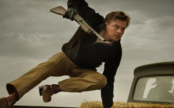 High-definition desktop wallpaper featuring a dynamic scene from 'Once Upon A Time In Hollywood' with a male actor charging with a weapon beside a classic car under an overcast sky.