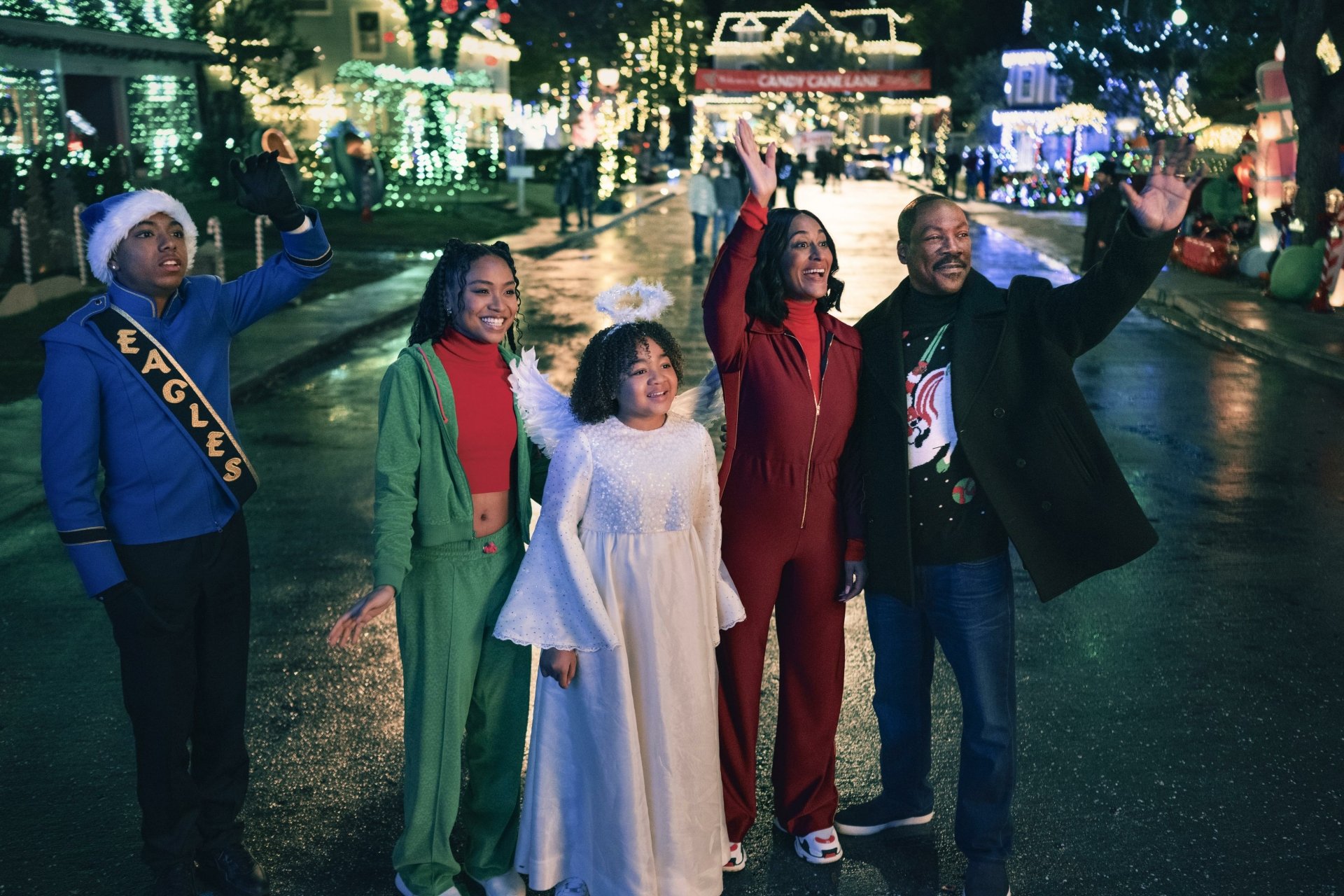 A festive scene from the movie Candy Cane Lane, featuring a group of five characters joyfully waving amidst a brightly decorated street illuminated with holiday lights.