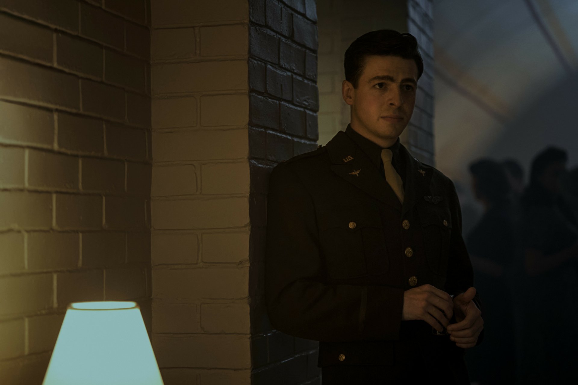 HD wallpaper featuring a scene from the TV show Masters of the Air, depicting a young man in a military uniform standing thoughtfully next to a lamp in a dimly lit hallway.