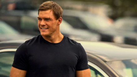 HD wallpaper featuring Alan Ritchson as the character Reacher from the TV show, standing confidently outdoors with cars in the background.