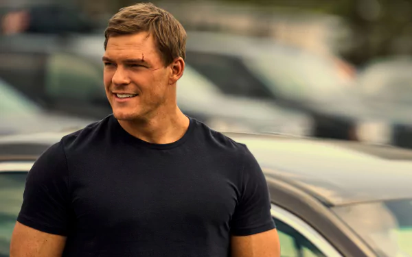 HD wallpaper featuring Alan Ritchson as the character Reacher from the TV show, standing confidently outdoors with cars in the background.