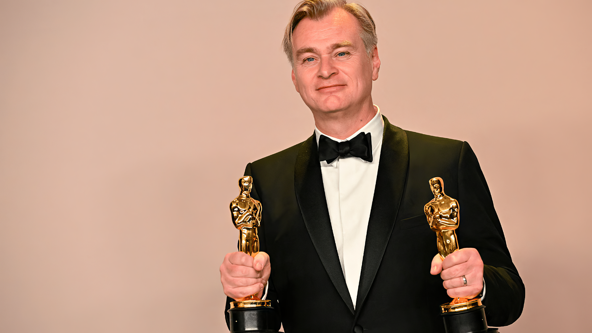 Christopher Nolan proudly holds two Academy Awards, dressed in a tuxedo, standing against a neutral background. A striking HD desktop wallpaper for film enthusiasts.