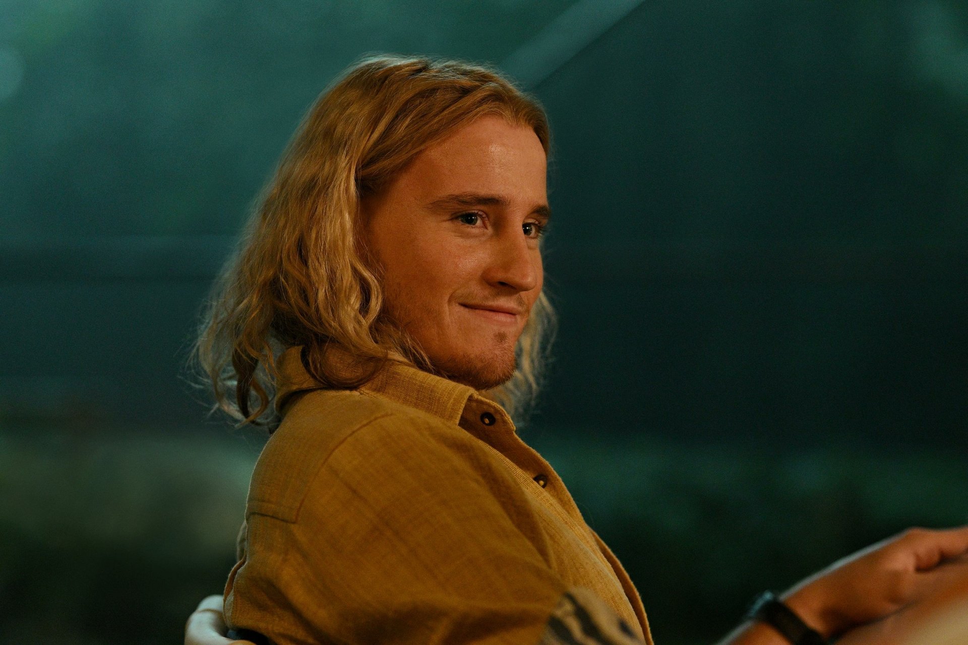 A character from the TV show Apples Never Fall smiles warmly, set against a softly lit background. This HD desktop wallpaper captures a moment of charm and intrigue.