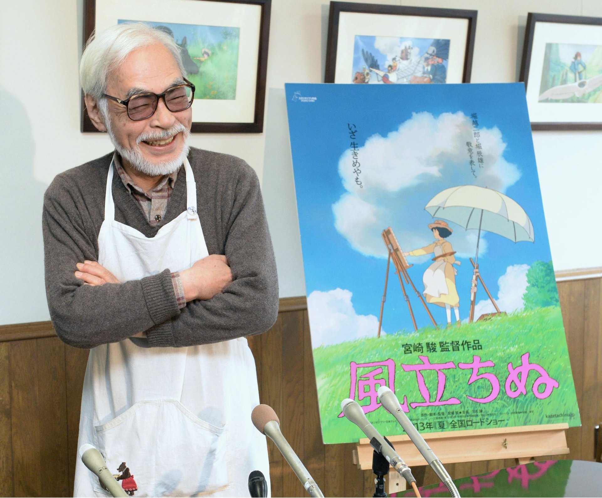 Hayao Miyazaki smiles beside a poster for his film, featuring a character with an umbrella in a vibrant, whimsical setting. A beautiful HD desktop wallpaper capturing his artistry.