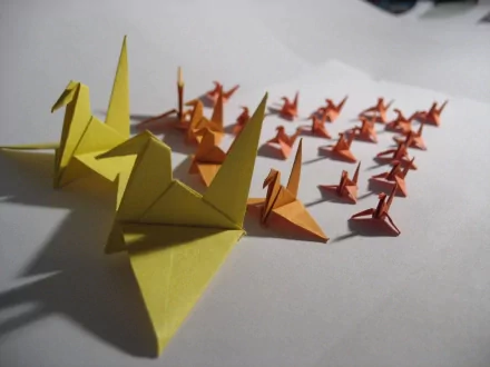 HD desktop wallpaper featuring a man-made origami display of multiple paper cranes in varying sizes and shades of yellow and orange on a white surface.