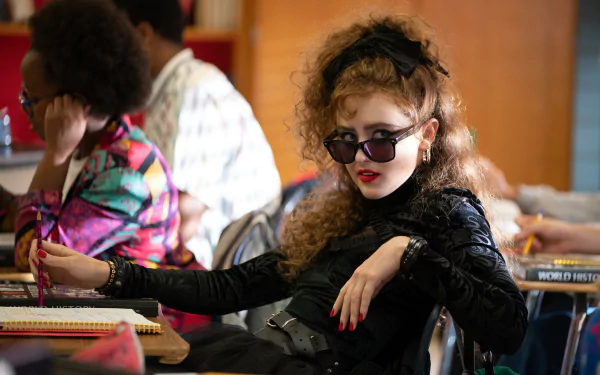 Stylish character from the movie Lisa Frankenstein sitting in a classroom, poised with sunglasses, portraying a bold and edgy look in a high-definition desktop wallpaper.