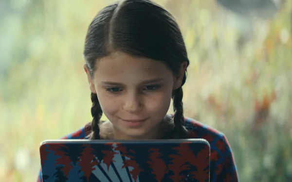 A young girl with braids looks intently at her colorful laptop in a serene outdoor setting. Background image from the TV show Constellation (2024), showcases intriguing storytelling.