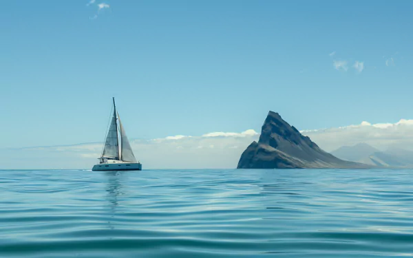 HD wallpaper featuring a catamaran sailing the ocean near an island with a prominent peak under a clear blue sky.