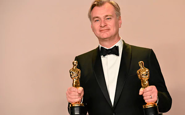 Christopher Nolan proudly holds two Academy Awards, dressed in a tuxedo, standing against a neutral background. A striking HD desktop wallpaper for film enthusiasts.