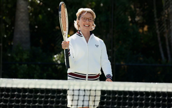 Smiling person with a tennis racket standing by the net on a sunny court, promoting the TV Show 'Apples Never Fall' as an HD desktop wallpaper.