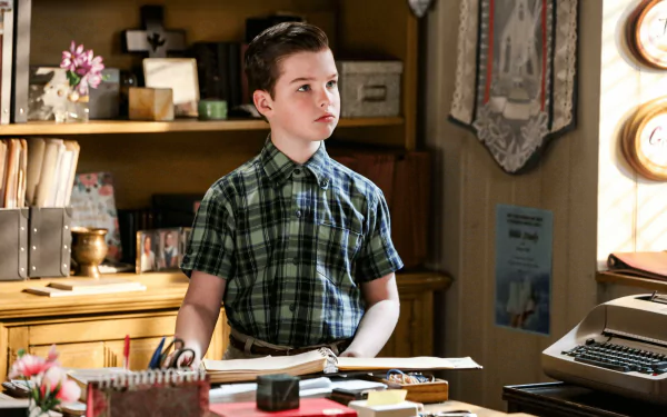 Young Sheldon character in a thoughtful pose at a desk with typewriter, HD wallpaper from the TV show.