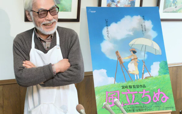 Hayao Miyazaki smiles beside a poster for his film, featuring a character with an umbrella in a vibrant, whimsical setting. A beautiful HD desktop wallpaper capturing his artistry.
