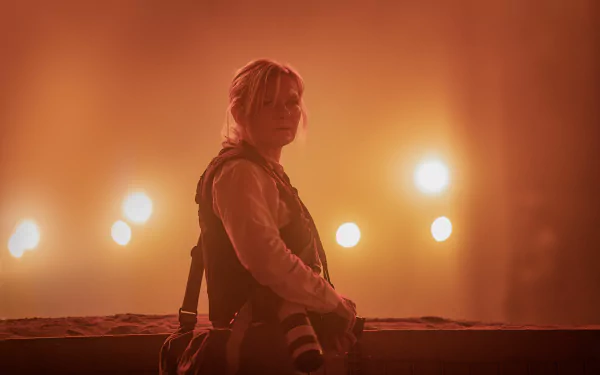 HD wallpaper featuring an individual resembling Kirsten Dunst from the side, set against a moody, amber-lit backdrop that creates a somber atmosphere, potentially from the movie Civil War (2024).