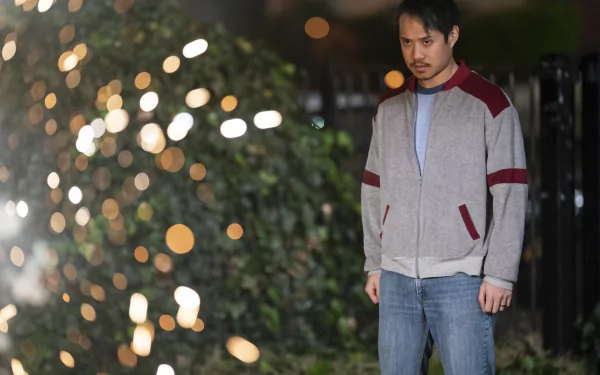 A man wearing a casual jacket stands in focus against a blurred background with sparkling lights, in a scene from the TV show The Sympathizer. The image is formatted as a high-definition desktop wallpaper.