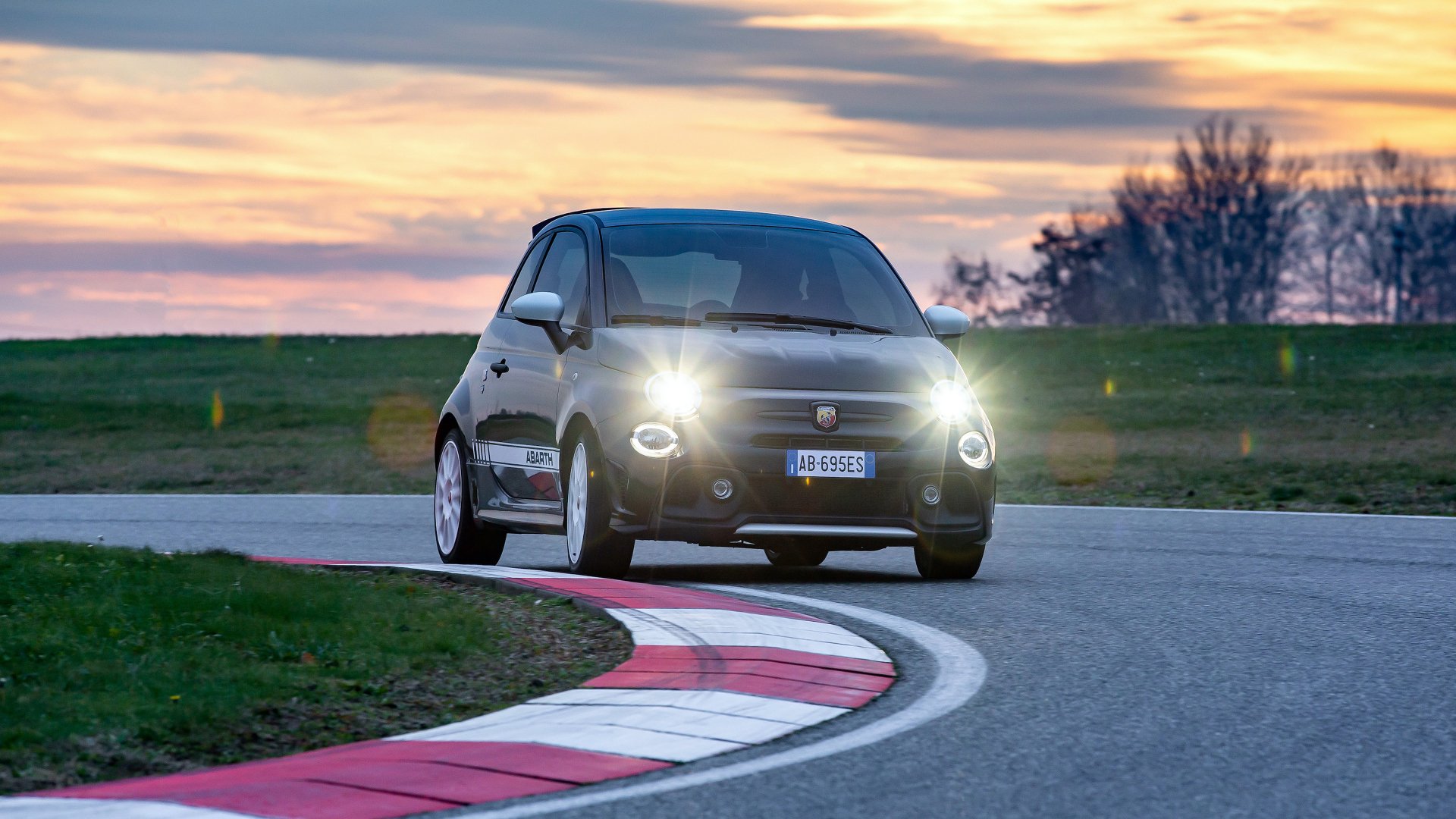 A black Fiat Abarth 695 Esseesse driving on a racetrack at sunset, with headlights on. The image is a high-definition desktop wallpaper and background.