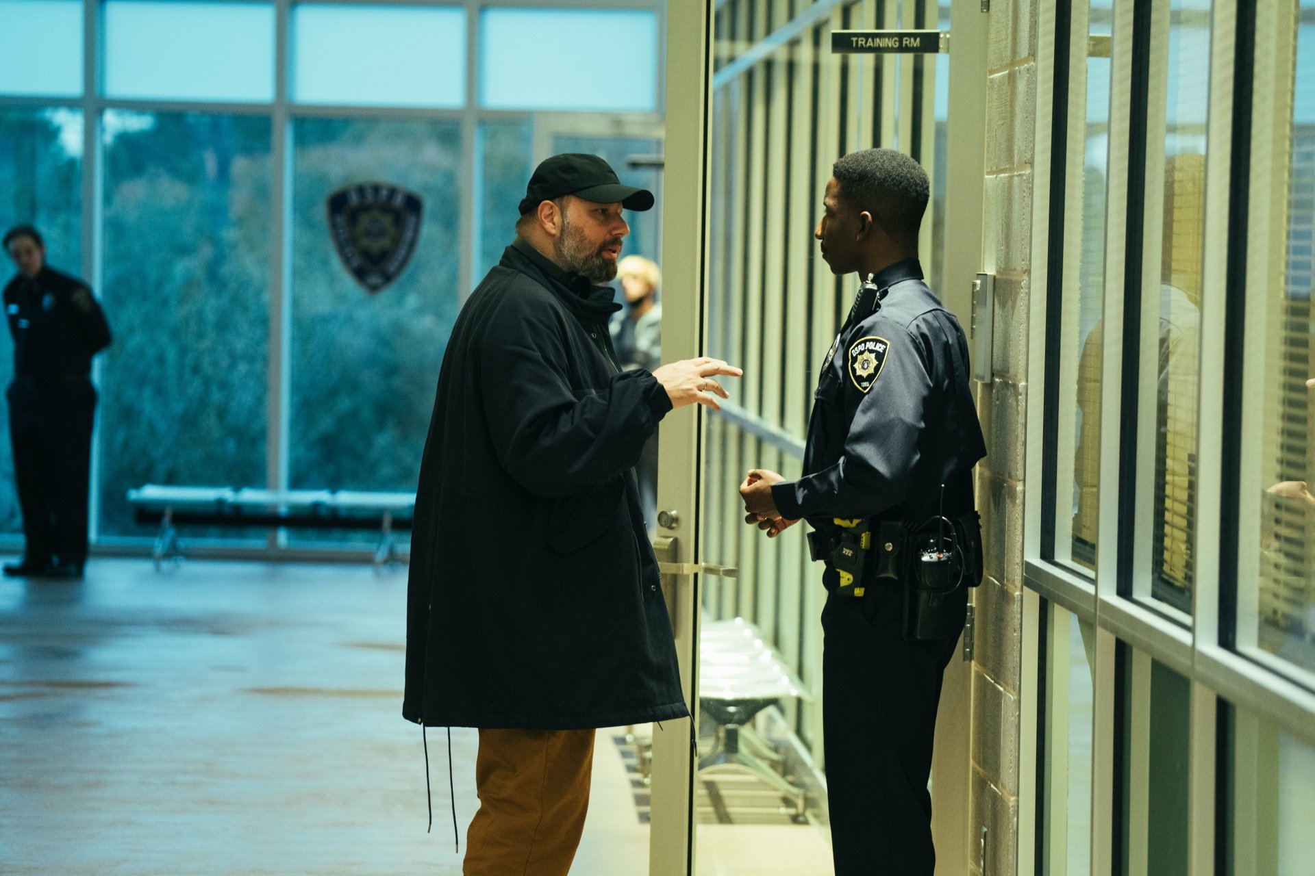 HD desktop wallpaper and background featuring a scene from the movie Kinds of Kindness, showing a man in a cap speaking with a uniformed officer in a well-lit indoor area.