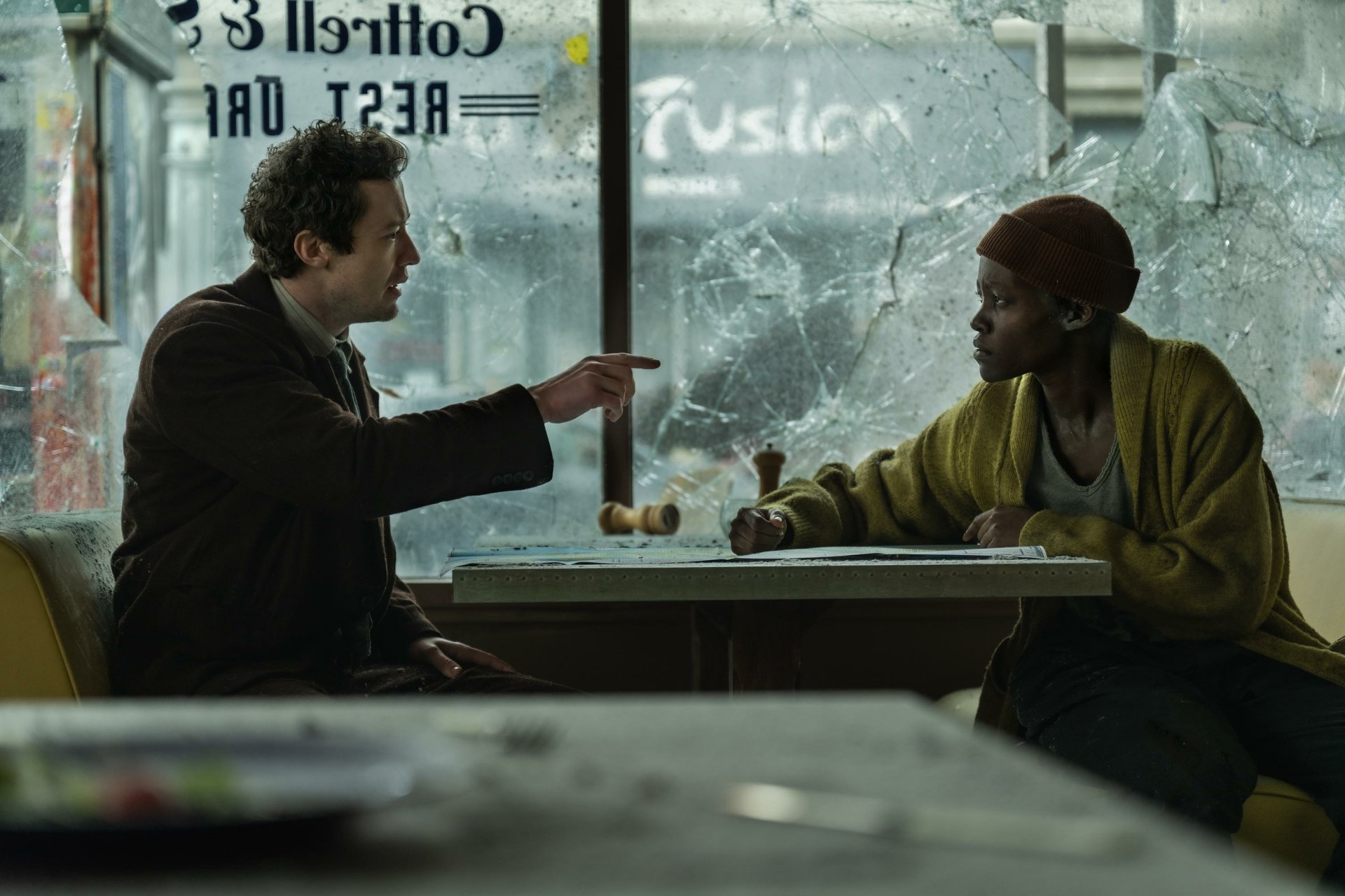 HD wallpaper from the movie A Quiet Place: Day One featuring Lupita Nyong'o and Joseph Quinn in an intense scene in a diner with broken windows.