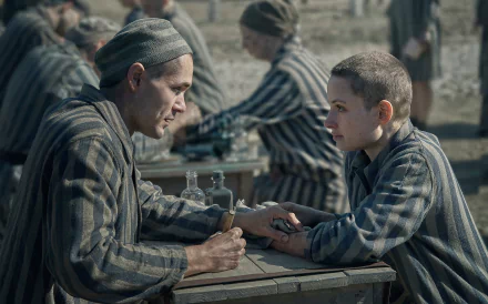 HD desktop wallpaper from the TV show The Tattooist of Auschwitz, featuring two characters in striped prison uniforms having a serious conversation at a table.