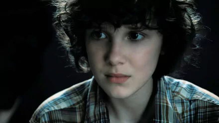 Close-up 8K Ultra HD wallpaper of Eleven from Stranger Things, portrayed by Millie Bobby Brown, featuring her intense and thoughtful expression in a dark setting.